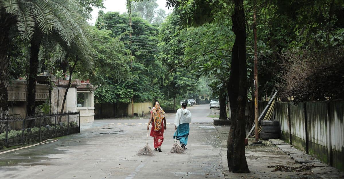 Two women sweeping a quiet street in Raipur, India, surrounded by lush greenery.