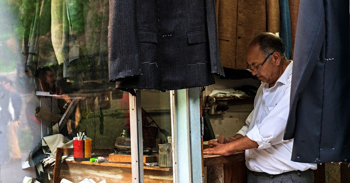 An experienced tailor working in a classic sewing shop surrounded by suits and materials.