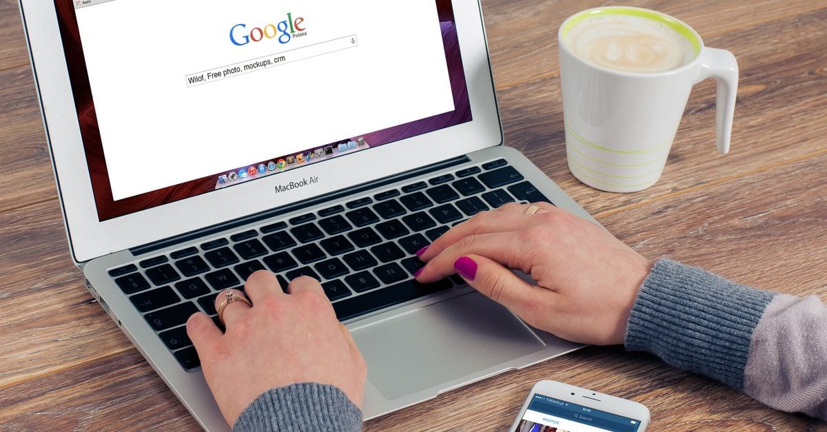 Hands typing on MacBook Air with Google search open, coffee nearby.