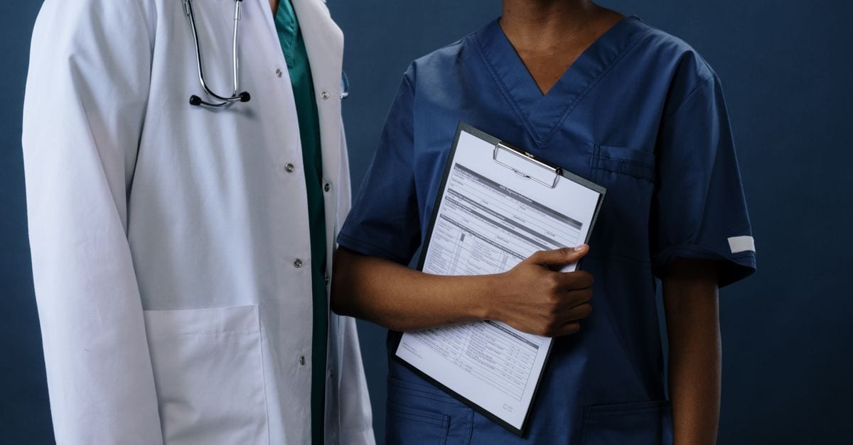 Two healthcare professionals in scrubs and lab coat with a clipboard, representing teamwork.