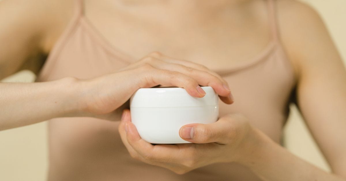 Close-up of a woman applying moisturizer with a focus on her hands and the cream container.