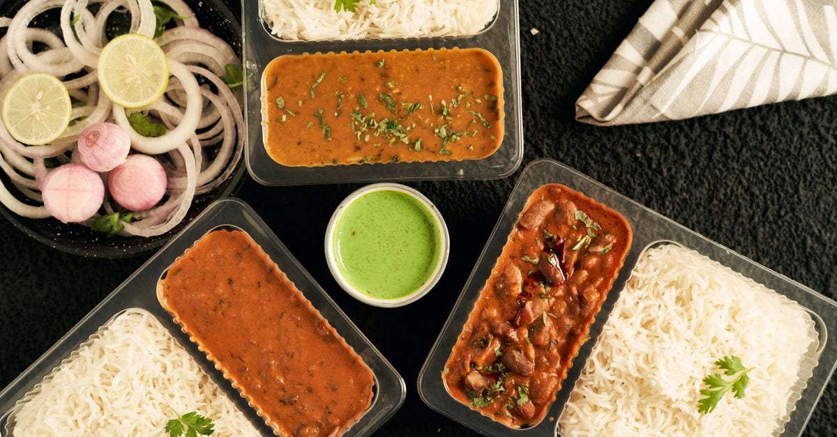 Top view of Indian curry, rice, and greens in takeaway containers with sliced onions.