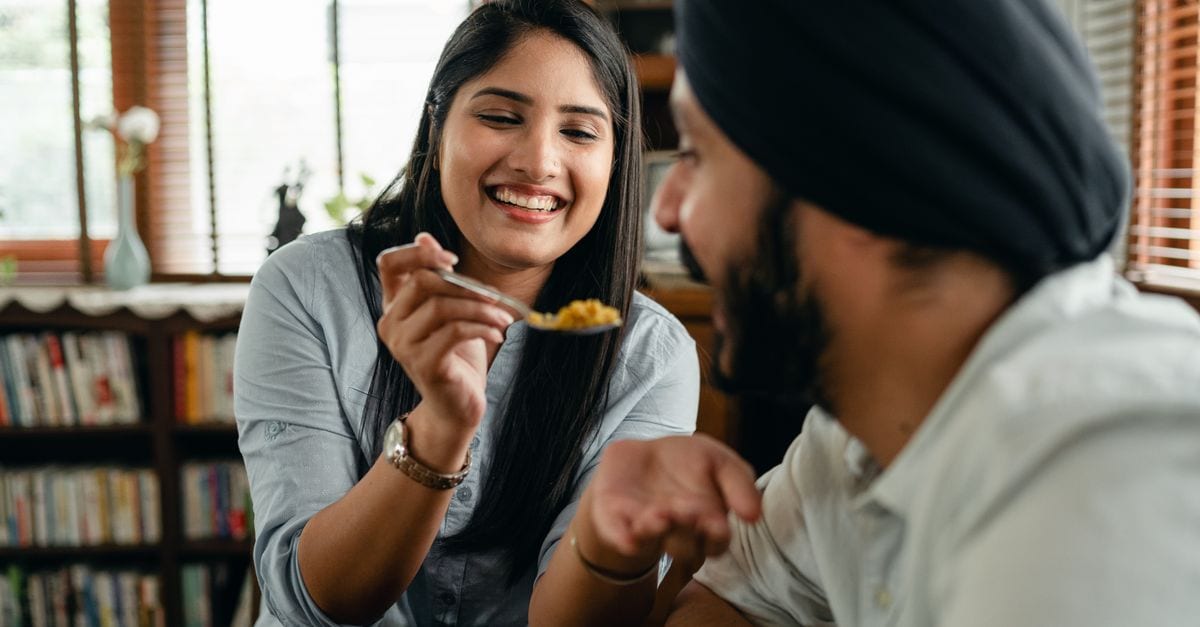 Happy young Indian woman smiling and feeding positive hungry boyfriend with delicious saffron rice